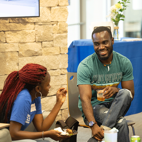  a Black male student wearing a green shirt smiling while talking to a black female student with red heard and a blue shirt in the welcome center. 