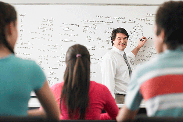 A white middle-aged male professor with short dark brown hair is standing inside a classroom in front of a whiteboard with mathematical equations written on the board in black. A black dry erase marker is visible in his right hand as he points to the board. He wears a white dress shirt, grey dress pants, and a grey necktie. He is smiling as he looks at the students sitting in the classroom. Their backs are to the camera. There are three students.