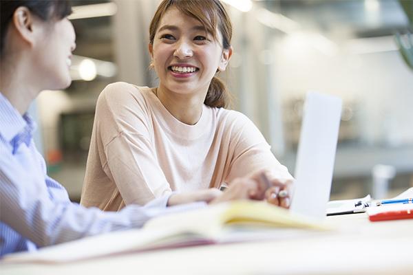 Two smiling female students of Asian heritage sit in a well-lit study area talking and laughing. The woman at left faces the camera and looks at her companion. She has medium-length brown hair in a ponytail and wears a light beige, long-sleeved shirt. Her friend has black hair and is looking away from the camera at the edge of the frame. She wears a blue-and-white pin-striped shirt, holds a pen, and has a laptop open before her. Books, pens, and notepads are visible but out of focus on a table.