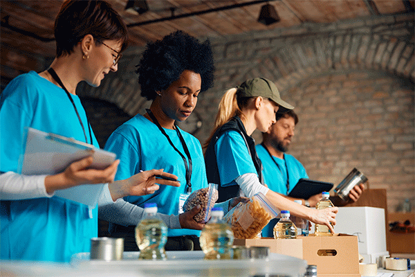 A diverse group of volunteers – three women and one man – work in a large room with brick walls to pack donated food items. They wear blue shirts and are placing Ziploc bags of food items, bottles of cooking oil, and cans of food into boxes. A smiling woman at the far right of the image holds a clipboard and appears to be directing the activity.